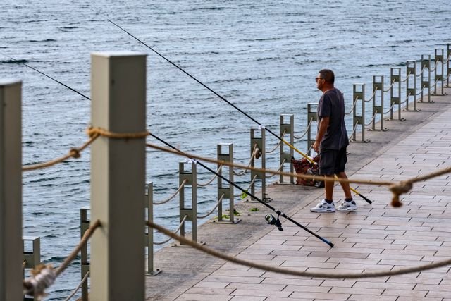 A man stands fishing with rods along a waterfront promenade, enjoying the peaceful day.